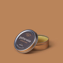 A round metal tin of Gatherwise All Heal Salve with its lid leaning against the open container, set against a solid warm brown background.