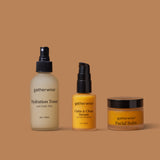 Three Gatherwise skincare products arranged on a warm tan background: the Hydration Toner and Daily Mist, a bottle of the Calm & Clear Serum, and a jar of theFacial Balm.