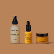Three Gatherwise skincare products arranged on a warm tan background: the Hydration Toner and Daily Mist, a bottle of the Calm & Clear Serum, and a jar of theFacial Balm.