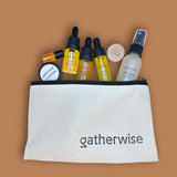 A cream-colored canvas cosmetic pouch with black trim and the Gatherwise brand name printed in black on the front, shown filled with various travel-sized skincare against a solid tan background.
